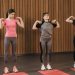 Three women in activewear stretching in an indoor yoga studio on red mats.